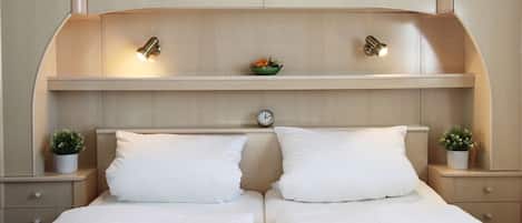 A double bed with white linens, a wooden headboard with built-in lighting, a clock, and two bedside tables with potted plants.