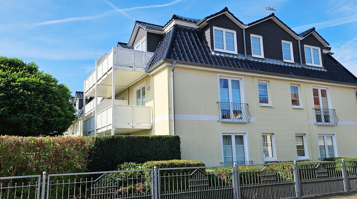 Ein mehrstöckiges Wohngebäude mit einem Balkon im zweiten Stock, einem dunklen Dach und einer beige farbenen Fassade.