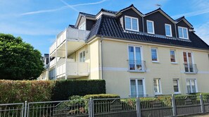 Un immeuble résidentiel à plusieurs étages, doté d’un balcon au deuxième étage, d’un toit foncé et d’une façade beige.