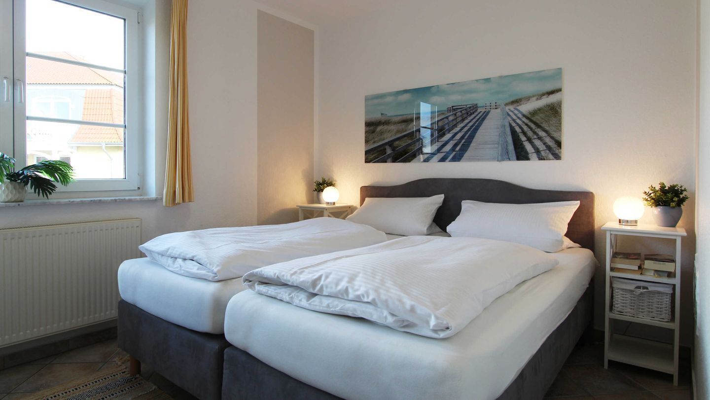 Ein Doppelbett mit weißer Bettwäsche, ein Nachttisch mit Lampe, ein Blumentopf und ein Gemälde mit Strandmotiv an der Wand.