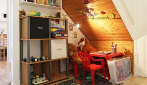 Ein gemütliches Dachzimmer mit Holzbalken, einem Bücherregal mit Spielzeug und Büchern, einem roten Sessel und einem Schreibtisch mit Lampe.