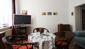 A living room with a television, wicker chairs, a table with a tablecloth, and artwork on the wall.