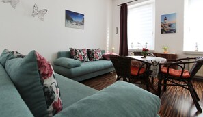 A living room with a teal sofa, a wicker chair, a small table with a vase, and a window with curtains.