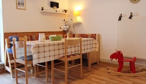 A cozy dining area with a wooden table set for two, a rocking horse, and a wall-mounted shelf with decorative items.