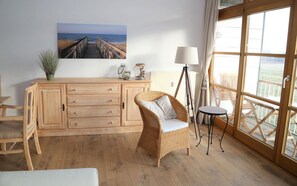 Ein Zimmer mit Holzboden, einem Korbstuhl, einem kleinen Tisch, einem Schrank, einer Lampe und einem großen Fenster mit Meerblick.