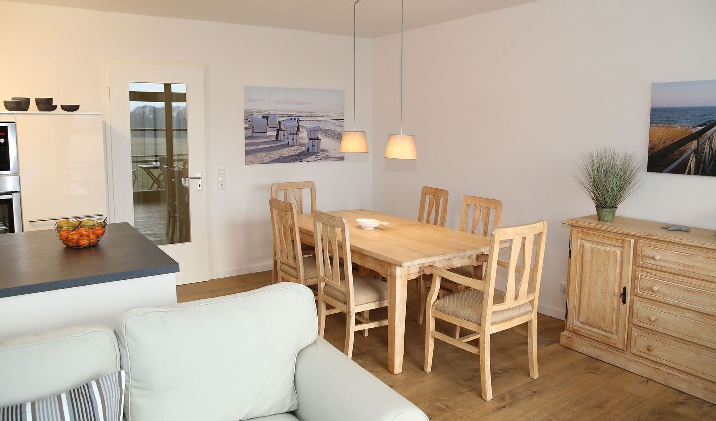 Ein Essbereich mit Holztisch und Stühlen, eine Küche mit Kühlschrank und Obstschale, und Blick auf den Strand durch eine Glastür.