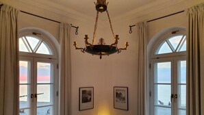 Una habitación con dos ventanas amplias, una araña y una mesa con lámpara.