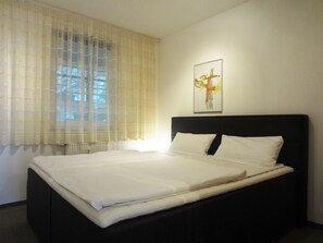 Una cama bien hecha en una habitación con un ventanal cubierto por cortinas translúcidas, un cuadro abstracto enmarcado en la pared y un radiador debajo de la ventana.