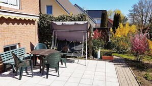 Une terrasse avec une table et des chaises, un espace salon abrité et un jardin agrémenté de plantes fleuries.