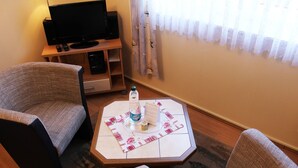 Une pièce accueillante avec deux fauteuils, une petite table octogonale surmontée d’un set de table à motifs floraux, une bouteille d’eau et un menu posé sur la table. Un téléviseur est installé sur un meuble en bois contre le mur, et une fenêtre avec des rideaux légers est visible.