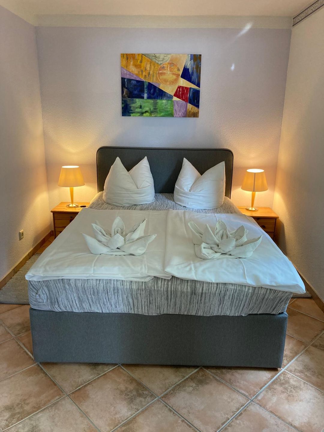 A neatly made bed with a grey headboard, two white pillows, and a decorative towel arrangement. There are two bedside lamps on wooden nightstands. A colorful abstract painting hangs above the bed.