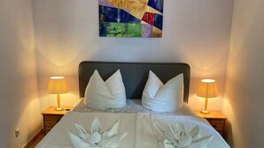 A neatly made bed with a grey headboard, two white pillows, and a decorative towel arrangement. There are two bedside lamps on wooden nightstands. A colorful abstract painting hangs above the bed.