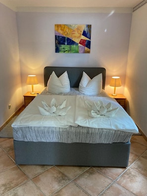 Una cama bien hecha con cabecera gris, dos almohadas blancas y un arreglo decorativo de toallas. Hay dos lámparas de noche de madera en las mesitas. Un cuadro abstracto colorido cuelga sobre la cama.