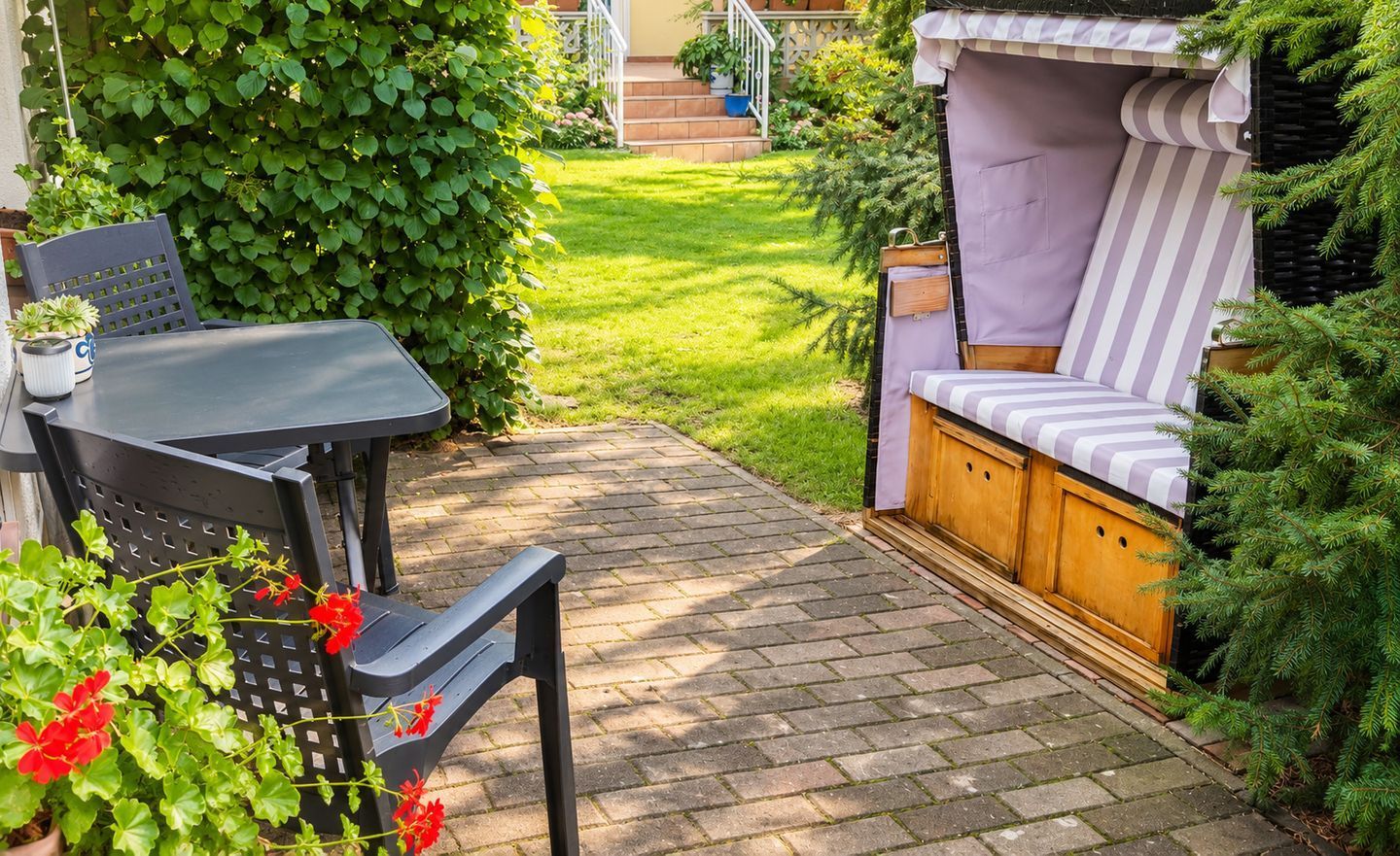Un patio con una mesa y sillas, un banco de madera y un pequeño jardín con seto y plantas en macetas.
