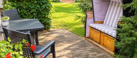 Un patio con una mesa y sillas, un banco de madera y un pequeño jardín con seto y plantas en macetas.