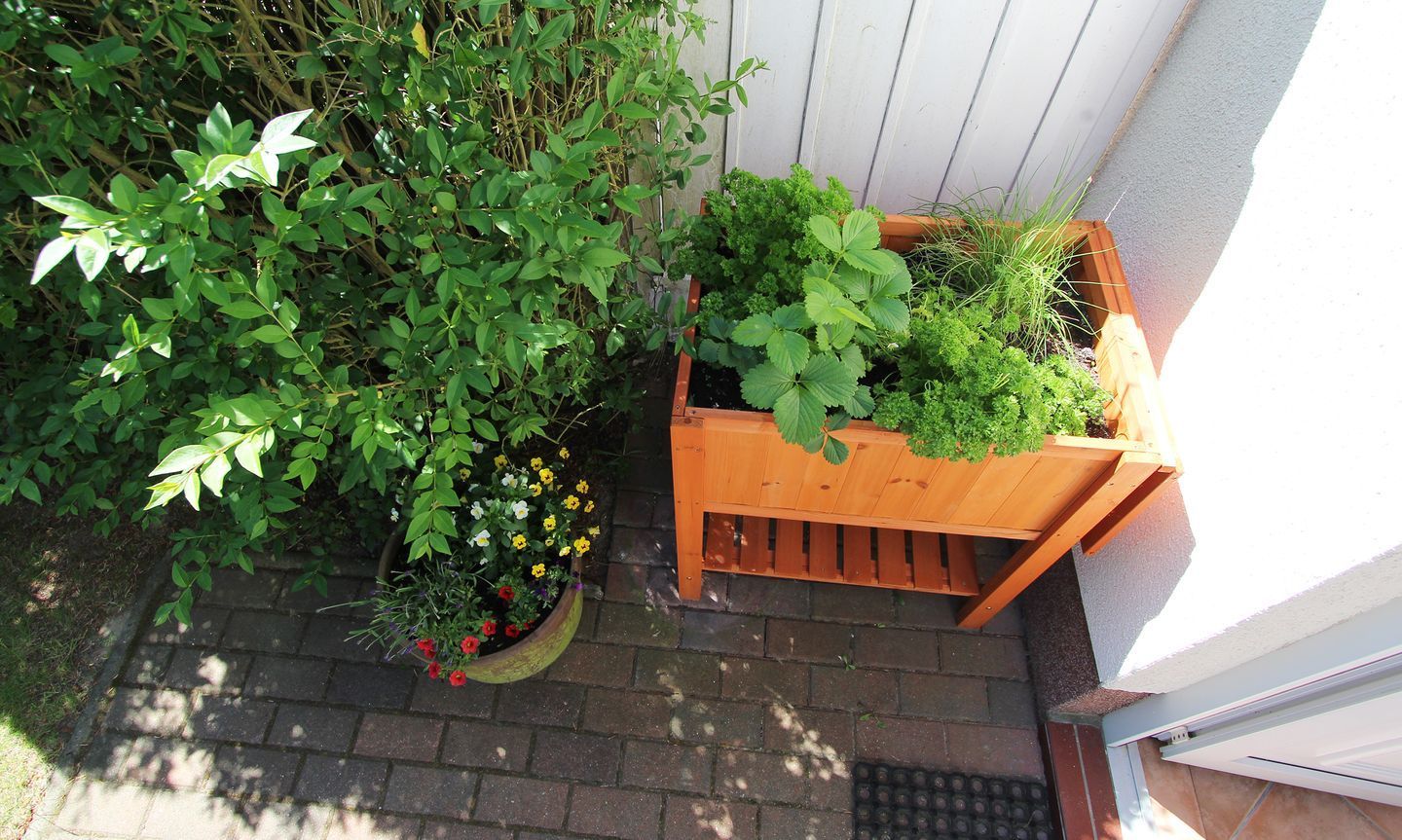 A wooden planter box with various plants and flowers, placed on a brick pathway next to a white wall.