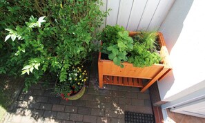 Une jardinière en bois, contenant diverses plantes et fleurs, est placée sur un chemin pavé de briques, à côté d’un mur blanc.