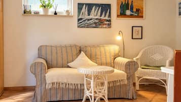 Un salón con un sofá a rayas, una mesa redonda de mimbre y una silla de mimbre. Hay cuadros en la pared y una ventana con persianas.