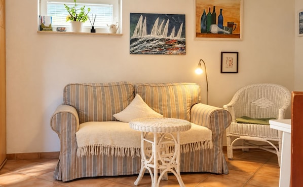 Un salón con un sofá a rayas, una mesa redonda de mimbre y una silla de mimbre. Hay cuadros en la pared y una ventana con persianas.