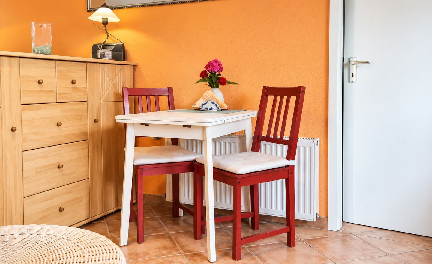 Une pièce avec un meuble en bois, une table blanche avec deux chaises, un vase de fleurs et un radiateur.
