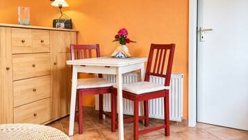 Une pièce avec un meuble en bois, une table blanche avec deux chaises, un vase de fleurs et un radiateur.