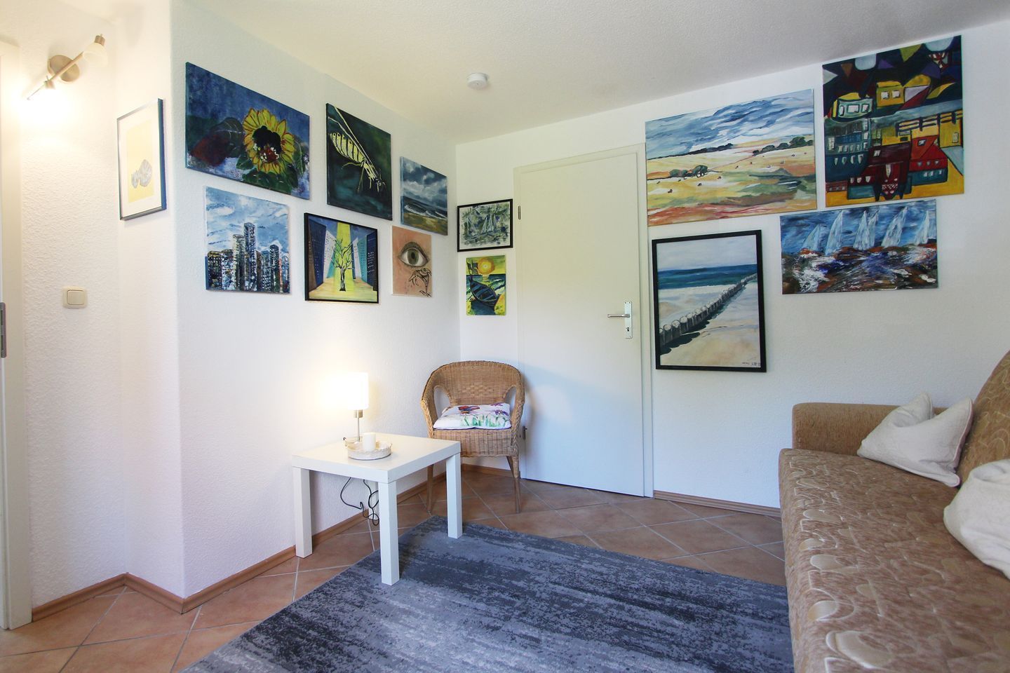 Een woonkamer met een bank, een stoel, een bijzettafeltje met een lamp en een vloerkleed. De muren zijn versierd met verschillende schilderijen.