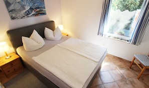 Una cama bien hecha con almohadas, una lámpara de noche y una ventana con vistas al verdor.