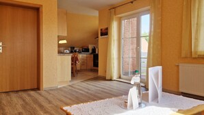 A room with a wooden door, a kitchen area with a microwave, a dining table with a lace tablecloth, and a bottle of water on the table. - Am Obstgarten - Comfortable vacation apartment with covered balcony near the beach in (Kühlungsborn)