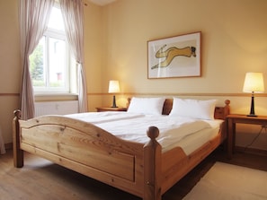 Una cama de madera con ropa de cama blanca, dos lámparas de noche, una ventana con cortinas y una obra de arte enmarcada en la pared.