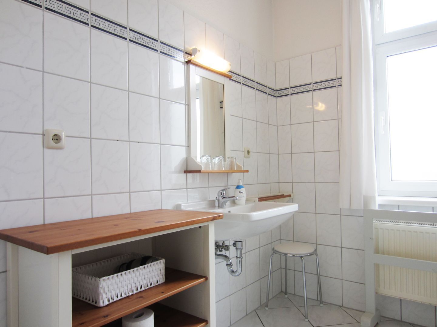 Et badeværelse med hvide flisemure, en træbordplade, en vask, et spejl, en skammel og en radiator.
