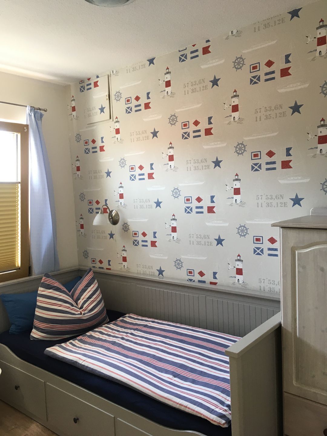 Une petite chambre à coucher accueillante, avec un lit simple, une table de chevet et un mur recouvert d’un papier peint sur le thème de la marine.