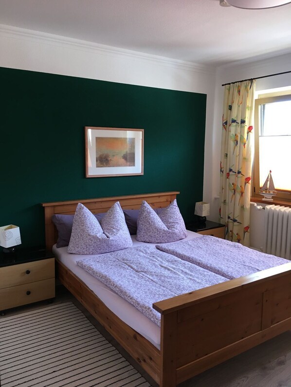A double bed with a wooden frame, a green wall, a framed picture, a nightstand with a lamp, and a window with curtains.