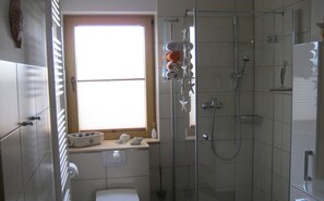 Un bagno con box doccia in vetro, una finestra con persiane, un portascopevoli con asciugamani e un lavandino bianco.