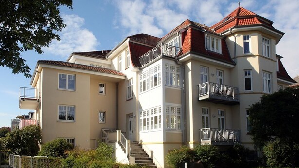 Ein mehrgeschossiges Wohngebäude mit einer zentralen Treppe, Balkonen und einem roten Ziegeldach.