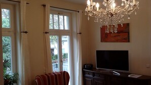 Ein Zimmer mit großem Fenster, einer Kristalllüster, einem Flachbildfernseher und einem abstrakten Kunstwerk an der Wand.