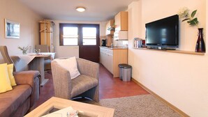 Un espacio habitable compacto con sofá, sillón, mesa de comedor, kitchenette y televisor montado en la pared.