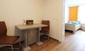 A compact room with a small table and two chairs, a bed with a blue mattress, and a window with orange curtains.