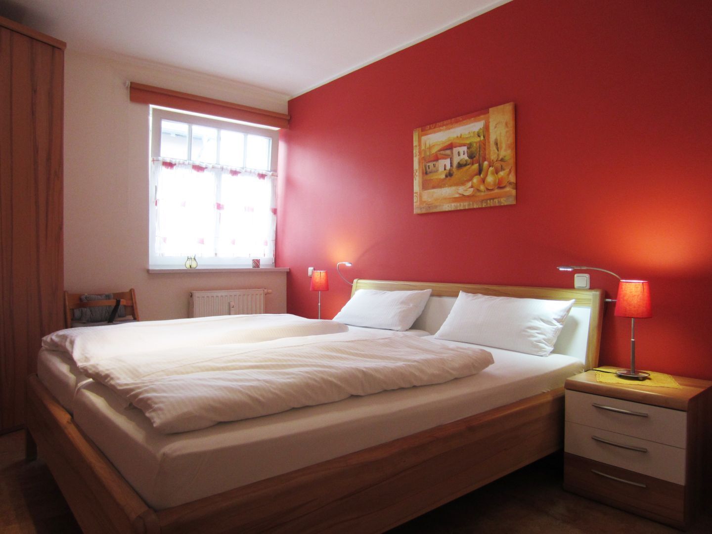 A neatly made bed in a room with red walls, a window with curtains, and a painting on the wall.