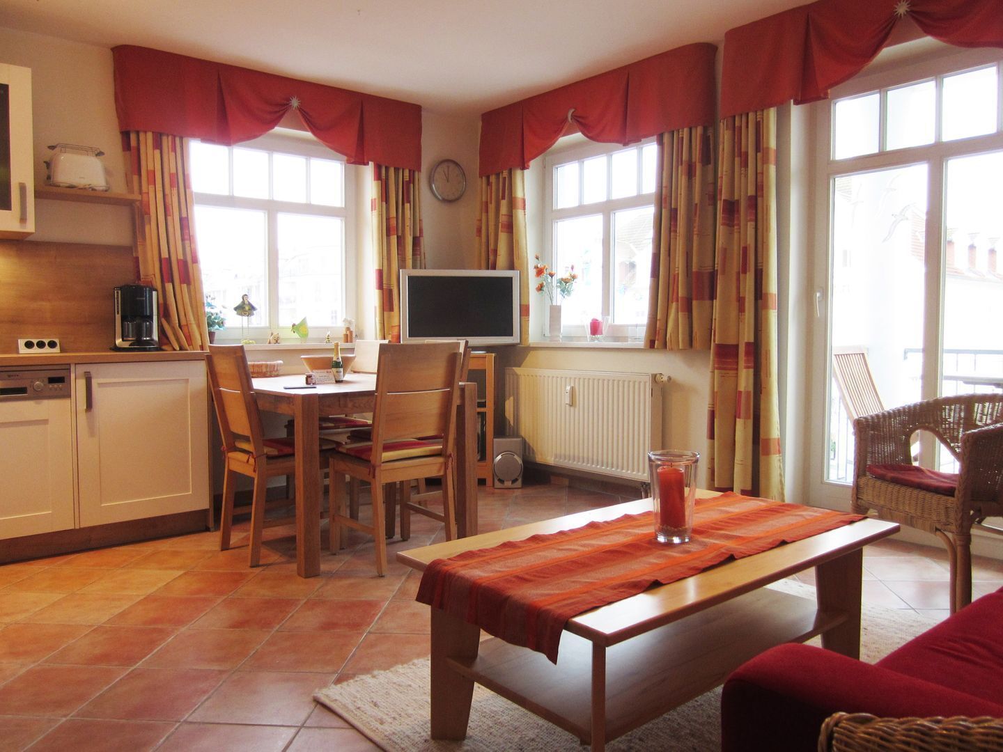 Ein gemütliches Wohnzimmer mit Holztisch, Stühlen, einer Couch und einem Fernseher. Es gibt eine Küchenzeile mit einer Kaffeemaschine und einer Uhr an der Wand.