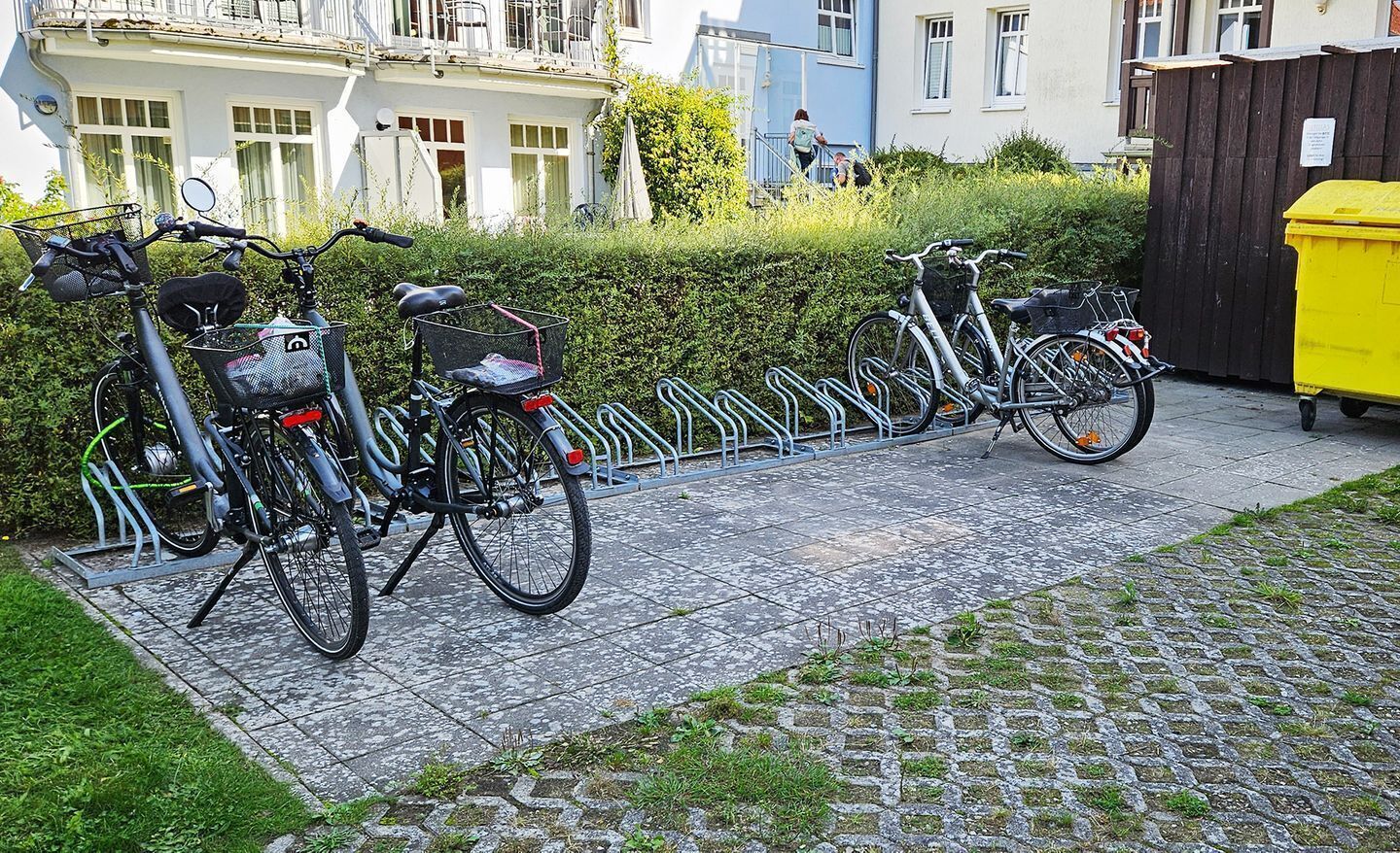 Eine Reihe Fahrräder, die in einem Fahrradständer an einem Kopfsteinpflasterweg in einer Wohngegend abgestellt sind, mit Häusern im Hintergrund.
