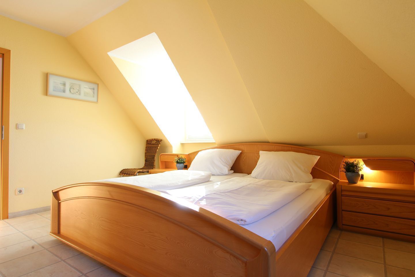 Ein Schlafzimmer mit einem Holzbett, einem Nachttisch, einem Korbstuhl und einem Oberlicht.