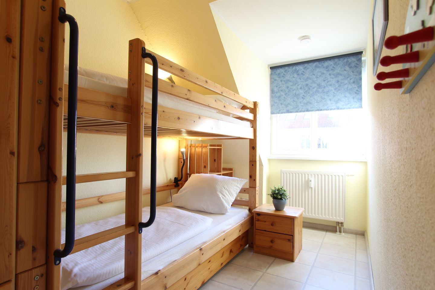 A bunk bed room with a wooden ladder, a bedside table with a plant, and a window with blinds.