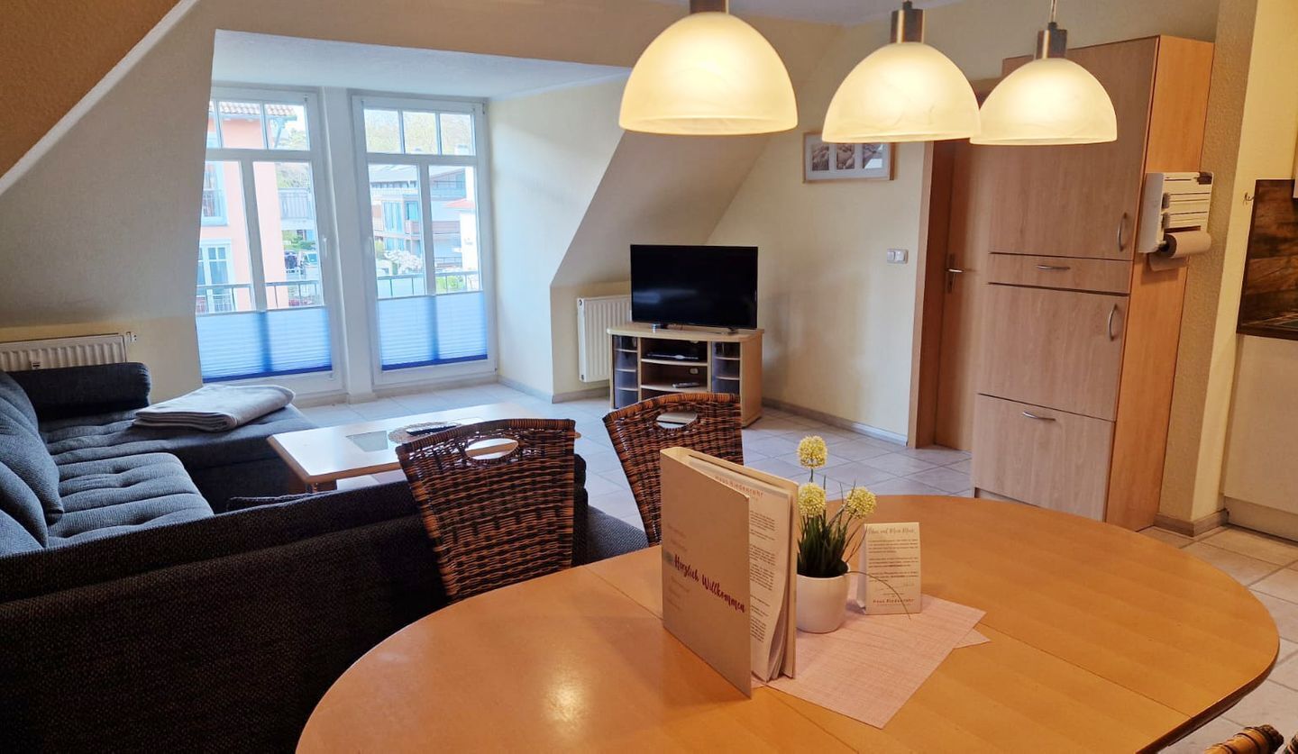 Ein modernes Wohnzimmer mit einer Couch, einem Esstisch, Korbstühlen, einem Fernseher und einer Küchenzeile mit Holzschränken und Pendelleuchten.