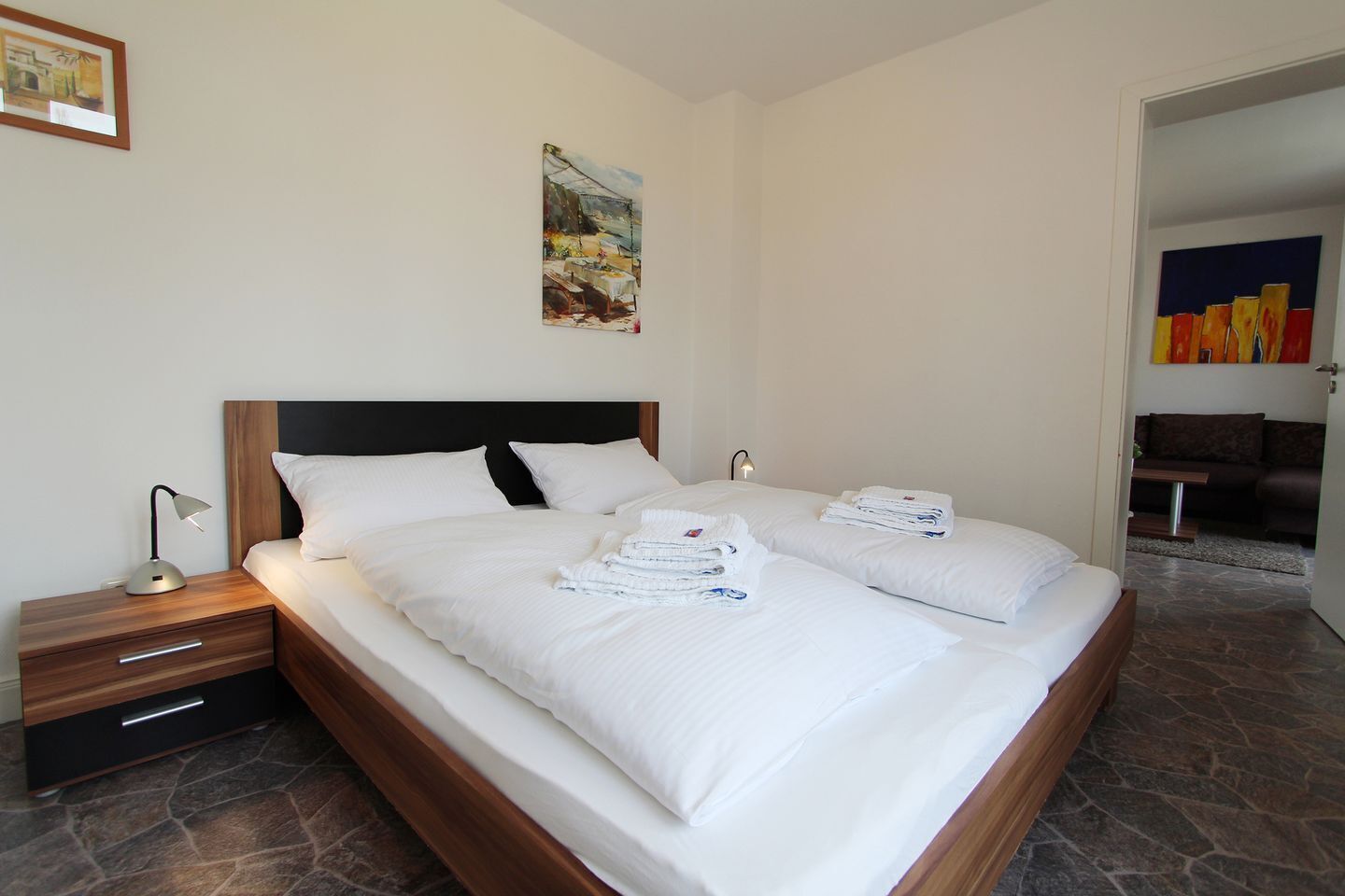 A double bed with white linens, a wooden headboard, a nightstand with a lamp, and a painting on the wall.
