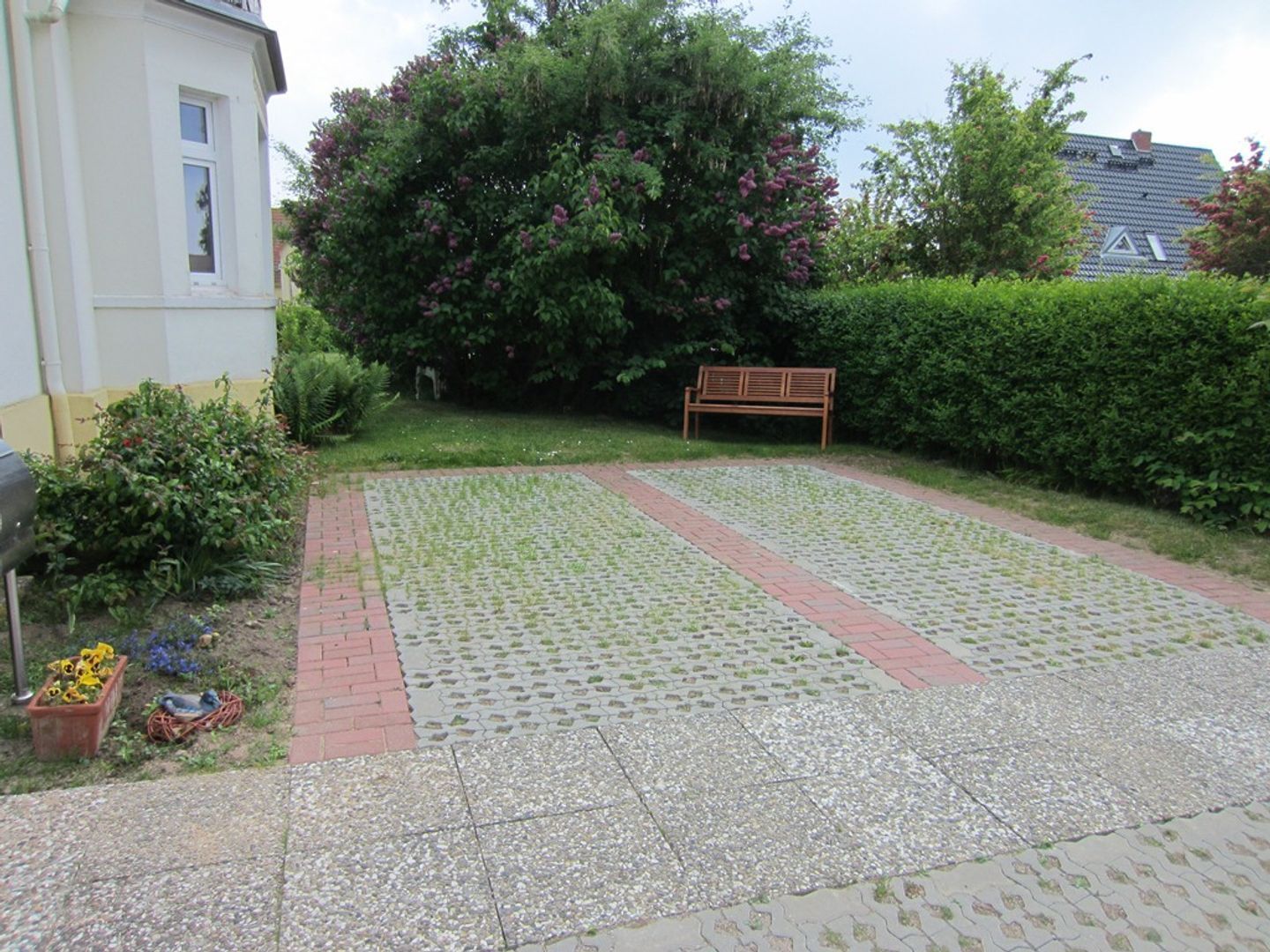 A paved patio with a bench, surrounded by a hedge and a large bush with pink flowers.