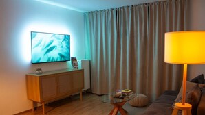 A living room with a flat-screen TV, a sofa, a lamp, a glass coffee table, and a radiator.