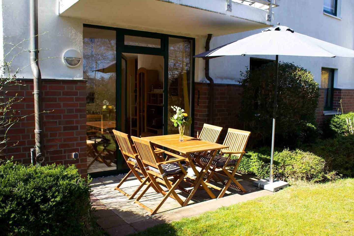 Een terras buiten met een houten tafel en stoelen, een plant in een pot en een witte parasol.