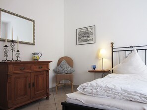Une chambre avec une commode en bois, un lit avec une literie blanche, une table de chevet avec une lampe, une chaise avec un coussin, un tableau encadré au mur et un miroir avec des bougies.