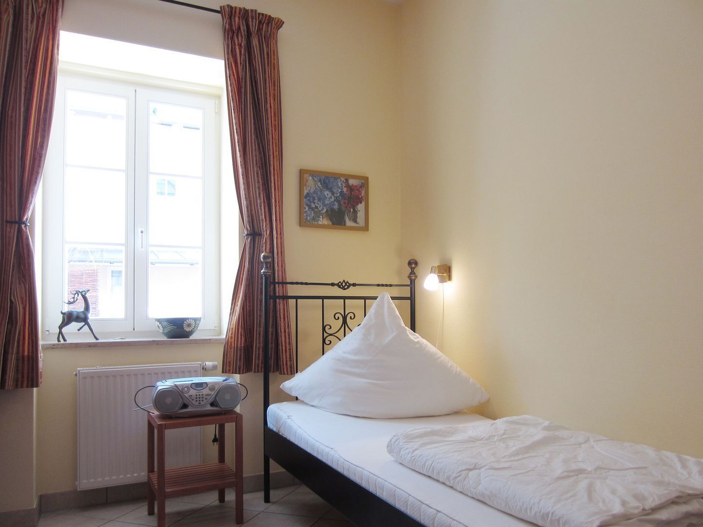 A small bedroom with a single bed, a radiator, a table with a radio, and a window with curtains.