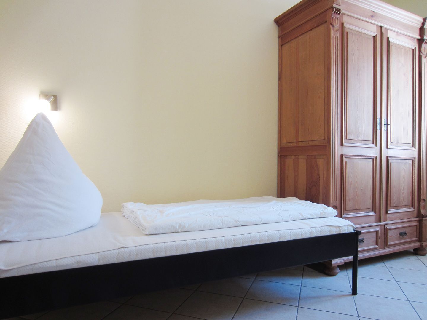 A single bed with a white mattress and pillow, a wooden wardrobe, and a wall-mounted light fixture.